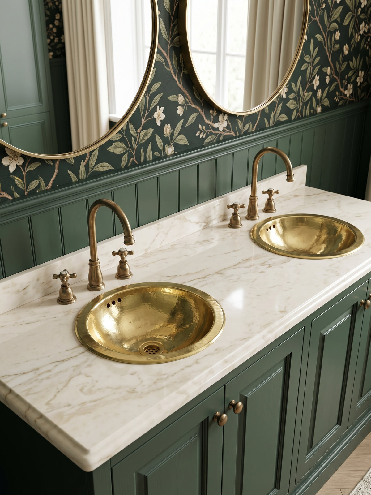 Two round hammered brass sinks in a white marble double vanity with green cabinets, brass faucets, floral wallpaper, and oval mirrors.