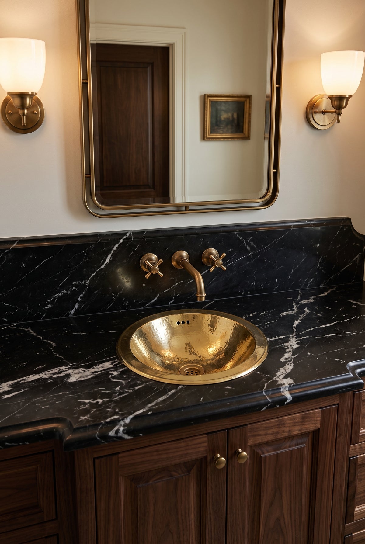 Round hammered brass sink in a black marble vanity beneath a brass wall-mounted faucet, brass-framed mirror, and sconces.