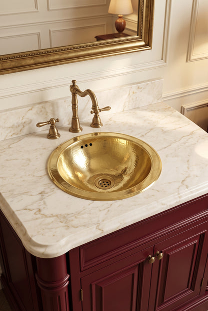 Round hammered brass sink in a cream marble vanity with a brass faucet, burgundy cabinets, and ornate gold-framed mirror.