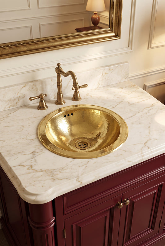 Round hammered brass sink in a cream marble vanity with a brass faucet, burgundy cabinets, and ornate gold-framed mirror.