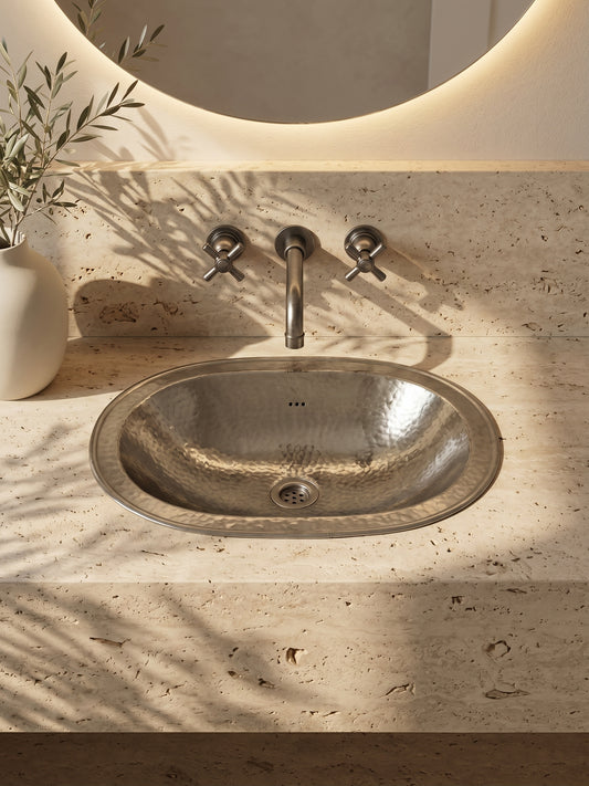 Oval hammered silver sink in a beige travertine vanity beneath a wall-mounted faucet and round backlit mirror.