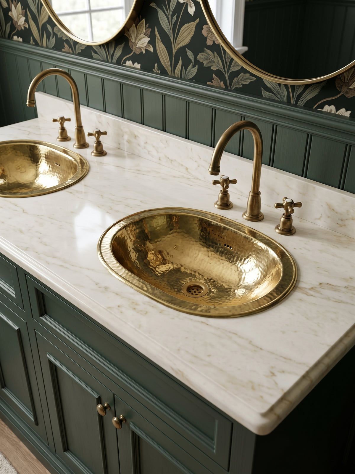 Two oval hammered brass sinks in a white marble double vanity with brass faucets, green cabinets, floral wallpaper, and oval mirrors.