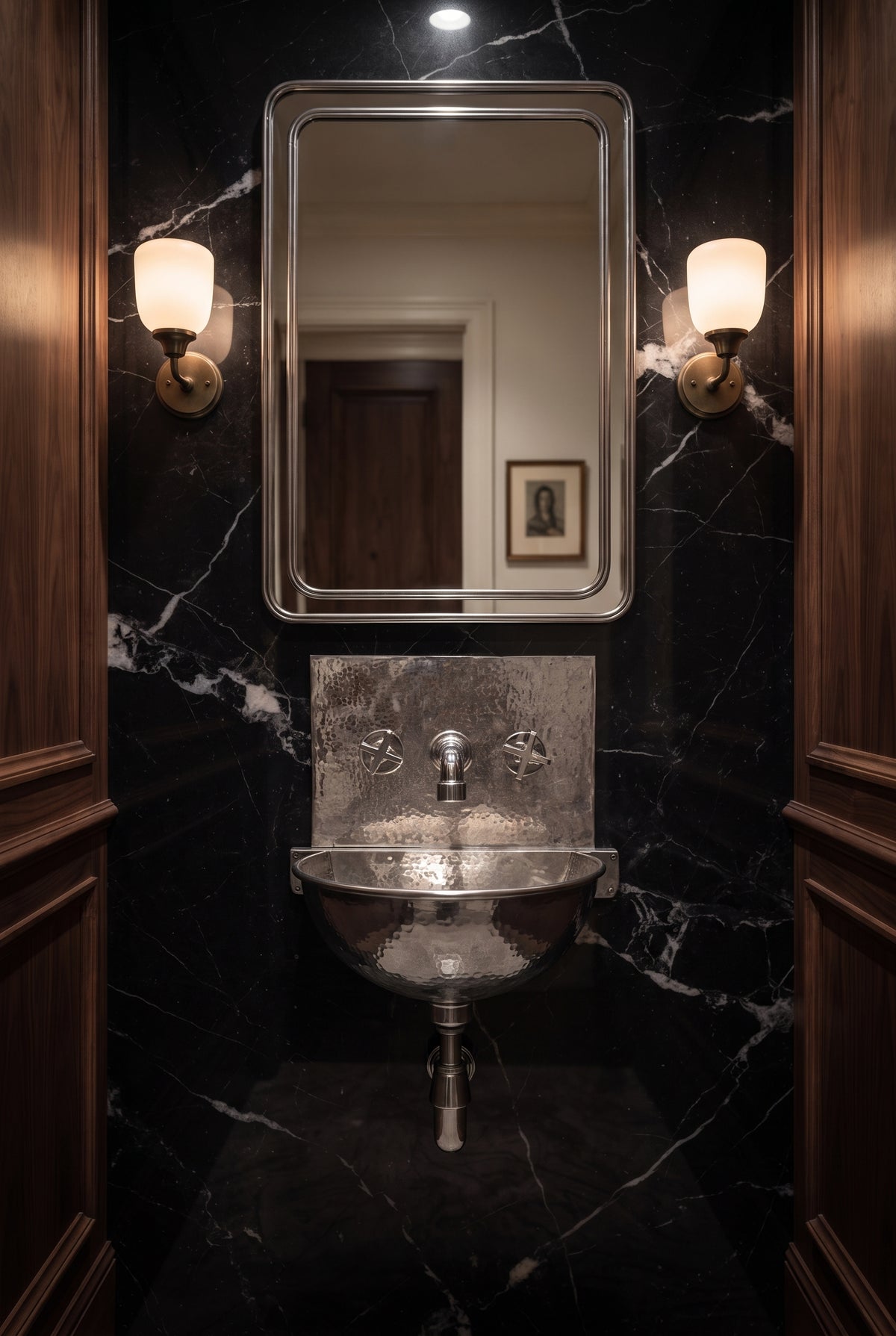 Hammered silver wall-mounted sink with matching faucet and exposed drainpipe against black marble, beneath a rectangular mirror and two sconces.