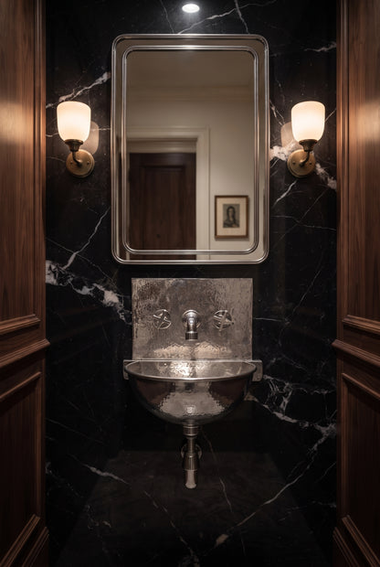 Hammered silver wall-mounted sink with matching faucet and exposed drainpipe against black marble, beneath a rectangular mirror and two sconces.