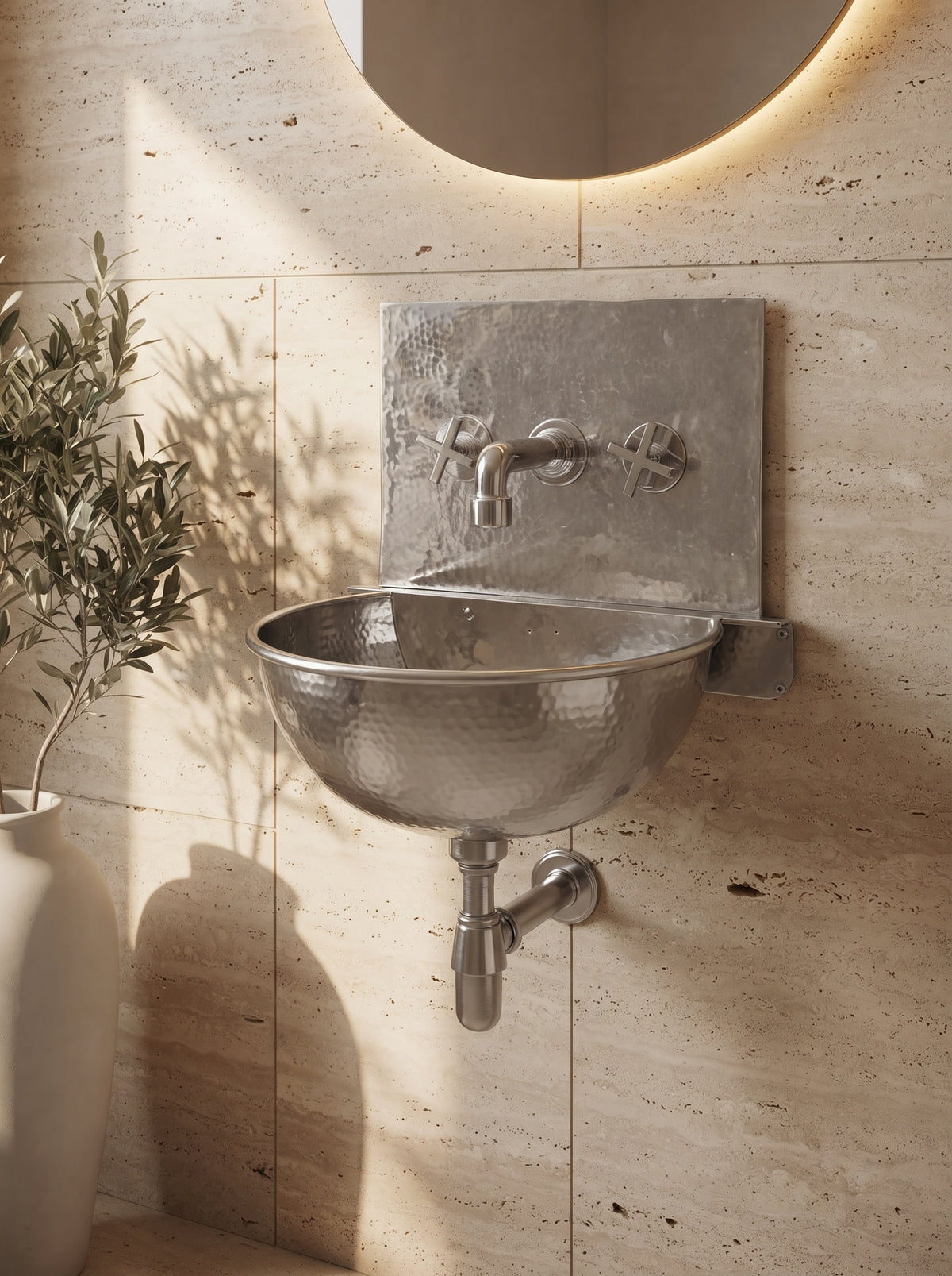 Close-up of a hammered silver wall-mounted sink and faucet against beige stone, with exposed plumbing, a round mirror, and a tall vase.