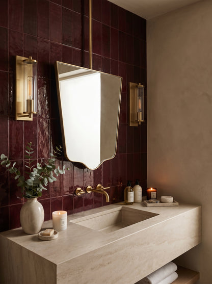 Geometric hanging brass mirror in a luxury bathroom with burgundy zellige tiles, a stone vanity, and brass fixtures.