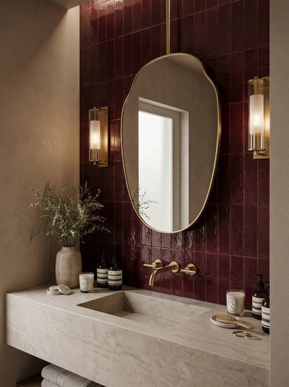 Luxury bathroom featuring a suspended organic brass mirror above a stone vanity with rich burgundy tiled walls.