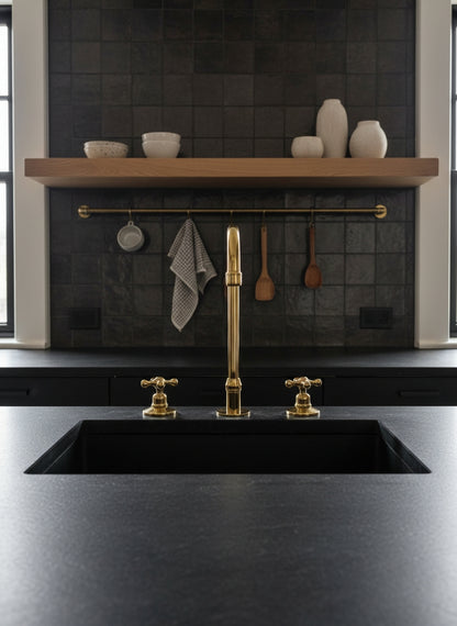 Unlacquered brass three-hole kitchen faucet with cross handles installed on a black stone countertop with dark tile backsplash.
