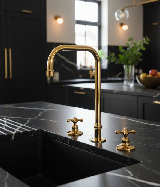 Unlacquered brass three-hole kitchen faucet with cross handles installed on a black marble kitchen island.