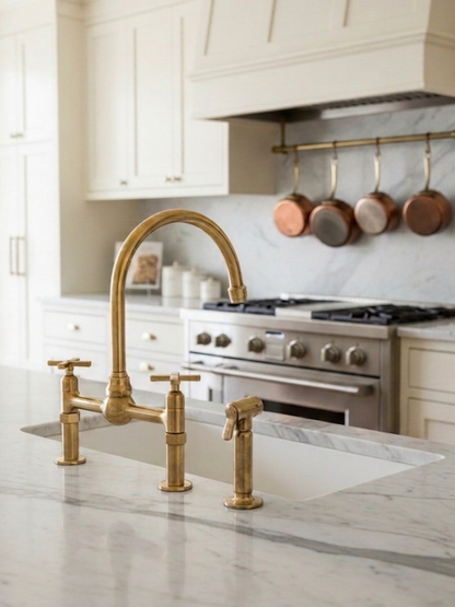 Unlacquered brass dual-handle bridge kitchen faucet with side sprayer installed on a white marble countertop in a bright luxury kitchen.