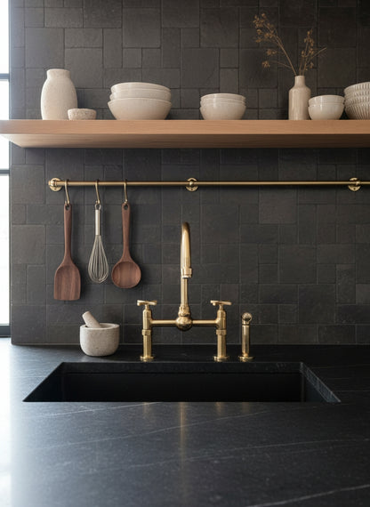 Unlacquered brass dual-handle bridge kitchen faucet with side sprayer installed on a black stone countertop beneath open wood shelving.