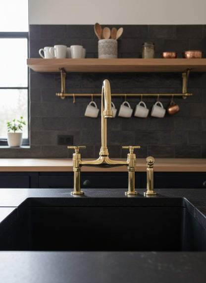 Unlacquered brass bridge kitchen faucet with side sprayer installed on a black countertop in a contemporary kitchen.