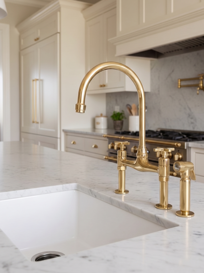 Unlacquered brass bridge kitchen faucet with side sprayer on a white marble countertop in a luxury cream kitchen.