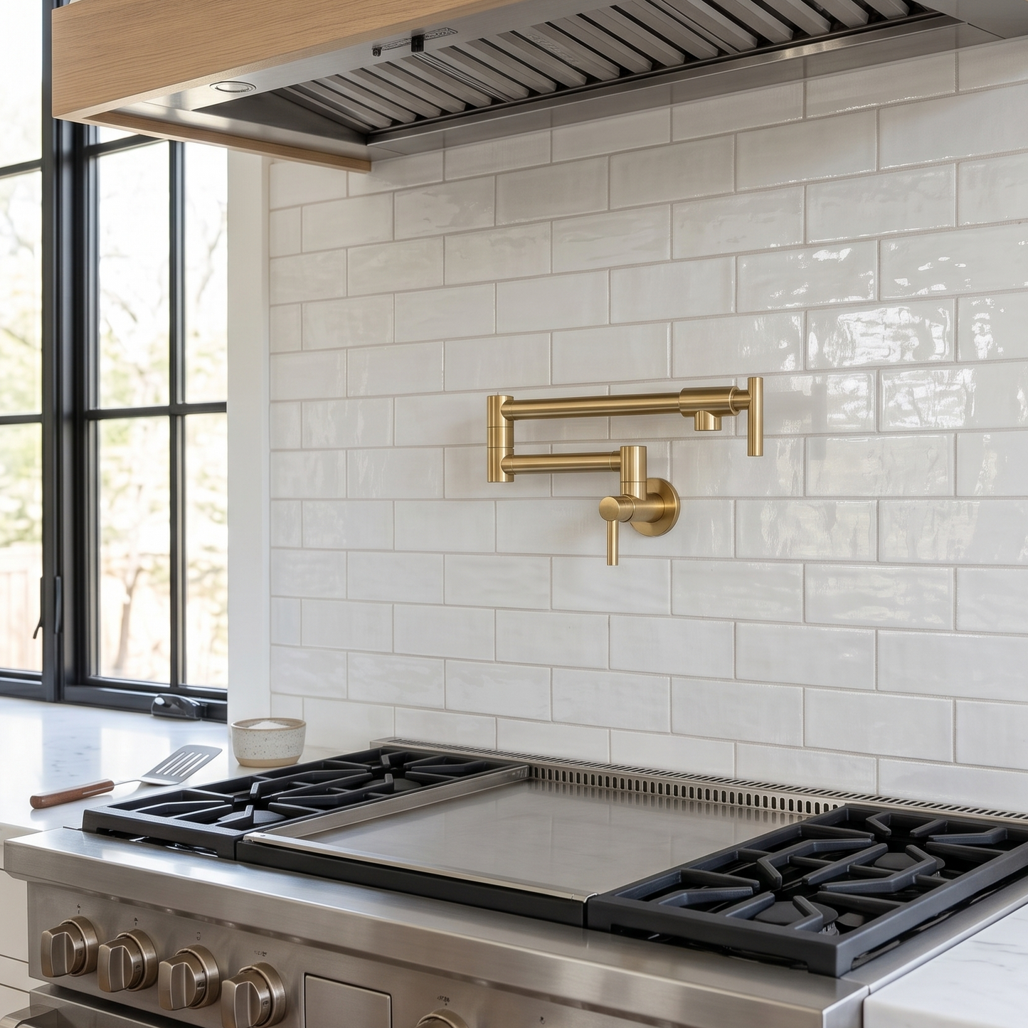 Unlacquered brass wall-mounted folding pot filler installed above a stainless steel range with glossy white subway tile backsplash and large black-framed window.