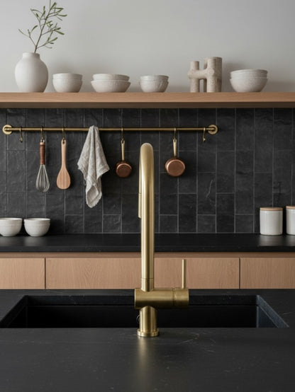 Unlacquered brass pull-down kitchen faucet installed on a black stone countertop with modern wood cabinetry and black tile backsplash.