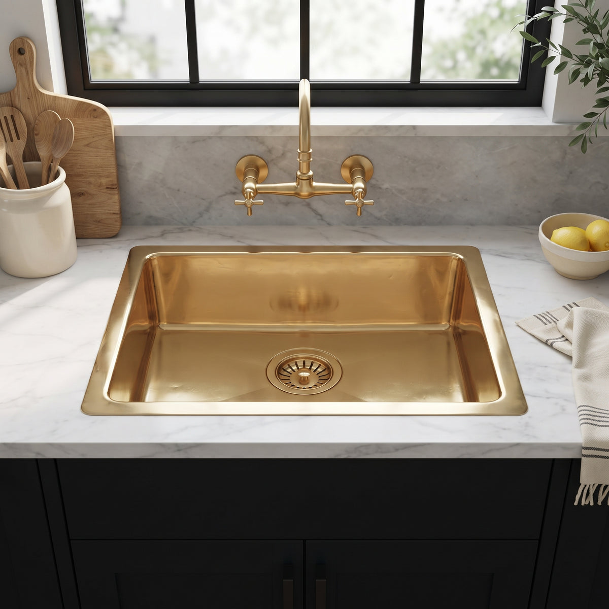 Brass drop-in kitchen sink beneath a wall-mounted brass faucet and black-framed window, set into white marble above black cabinets.