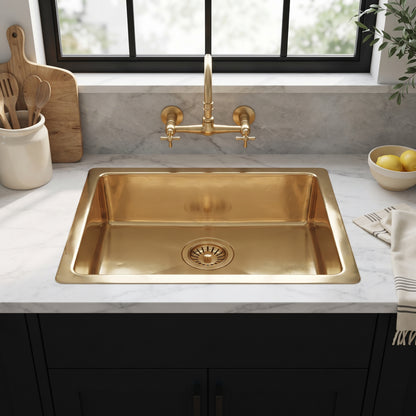 Brass drop-in kitchen sink beneath a wall-mounted brass faucet and black-framed window, set into white marble above black cabinets.