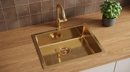 Brass drop-in sink with matching gooseneck faucet on an oak countertop, against brown tiles beside a basil plant.