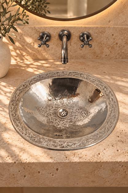 Oval silver sink with an engraved floral center and decorative rim in a travertine vanity beneath a wall-mounted faucet.