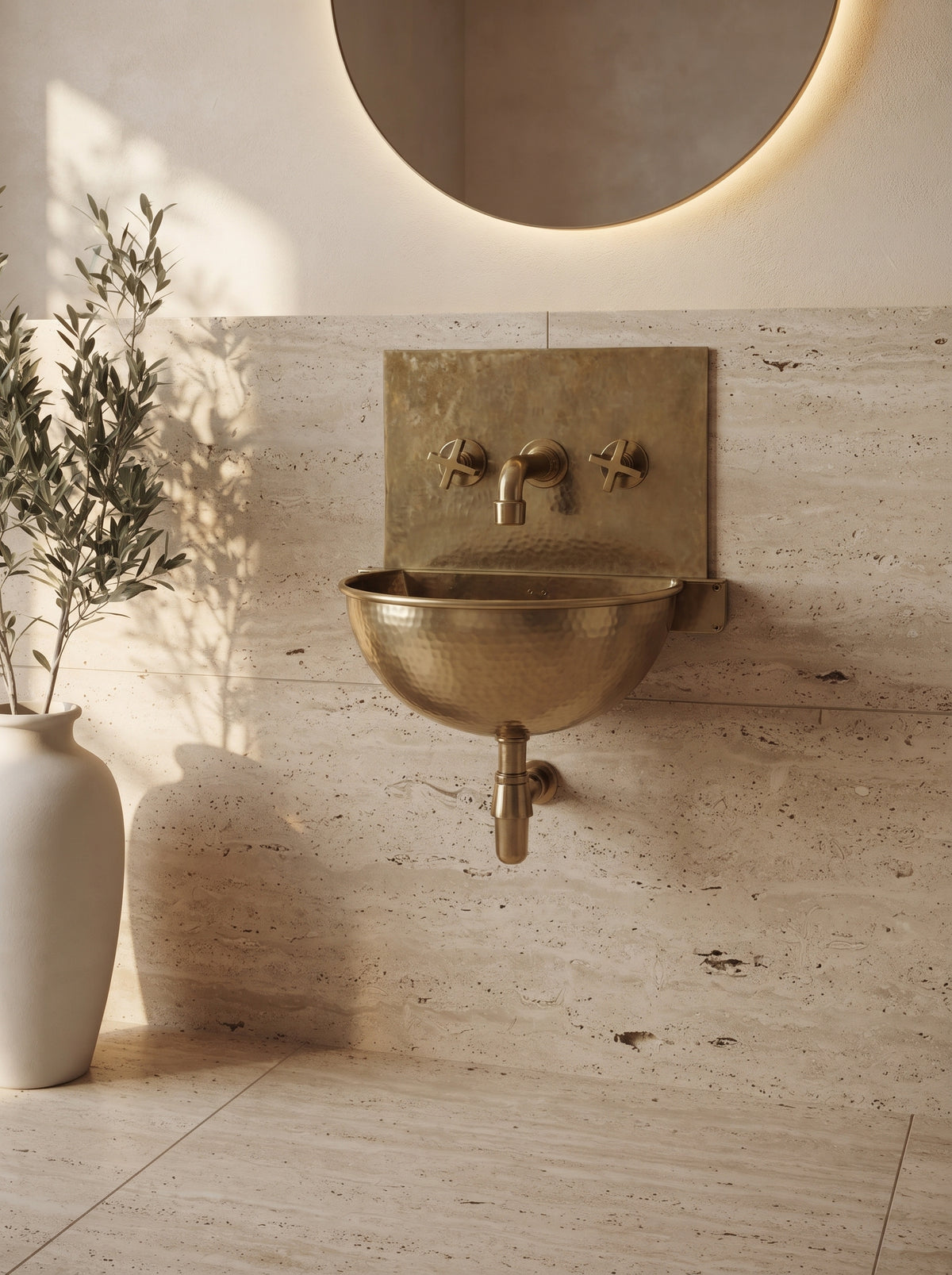 Unlacquered brass wall-mounted sink with matching backsplash and exposed drainpipe against beige stone, beneath a round mirror.