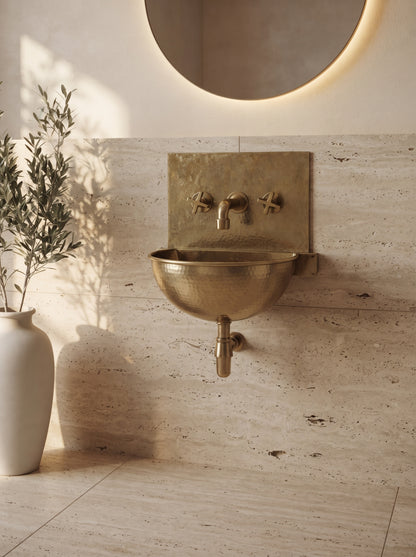 Unlacquered brass wall-mounted sink with matching backsplash and exposed drainpipe against beige stone, beneath a round mirror.