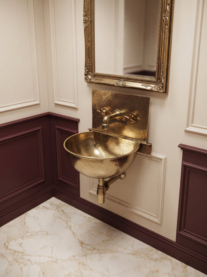 Unlacquered brass wall-mounted basin with integrated faucet beneath an ornate gold mirror, surrounded by cream and burgundy paneling.