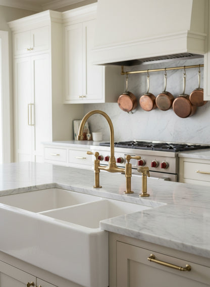 Unlacquered brass bridge kitchen faucet with side sprayer over a white farmhouse sink and marble countertop in a classic kitchen.