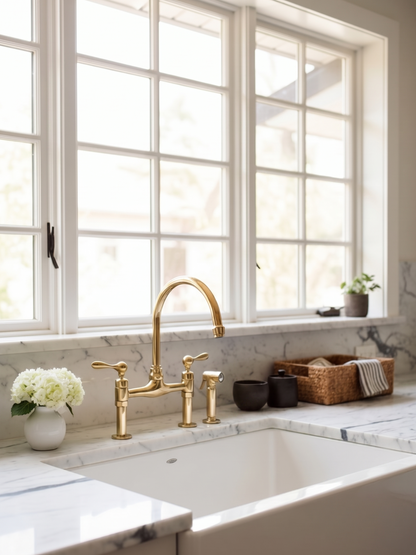 Vintage brass bridge faucet with side sprayer installed on a white marble countertop beneath large kitchen windows.