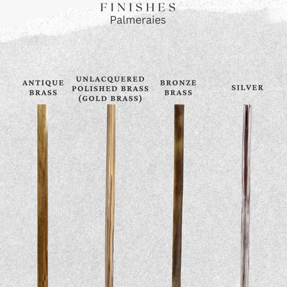 Four slender metal palmeraie stems in different finishes are displayed against a light textured background with labels: Antique Brass, Unlacquered Polished Brass (Gold Brass), Bronze Brass, and Silver.