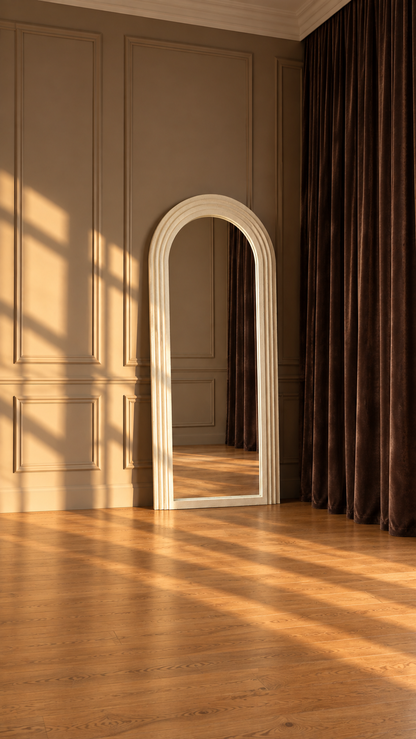 An arched white mirror with a double-trim frame stands on a polished wooden floor in a room with beige paneled walls and dark brown curtains, lit by warm sunlight casting diagonal shadows.