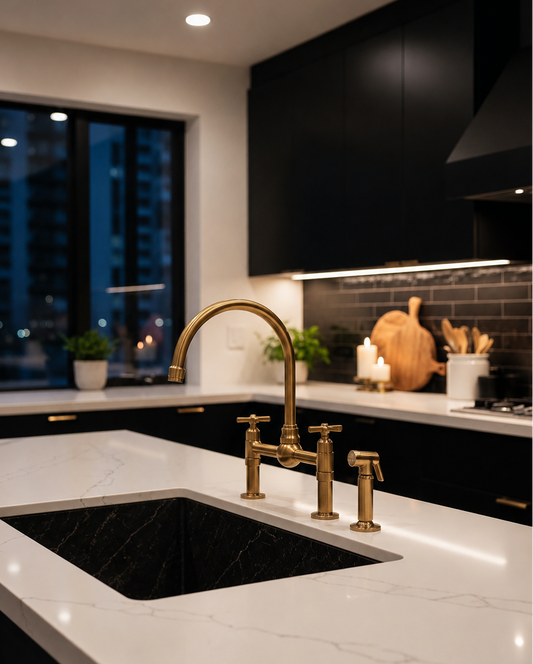 A modern kitchen scene with a white marble countertop, black sink, and a prominent brushed brass faucet and matching fixtures. Black cabinetry, a dark subway tile backsplash, and a window view at night are visible in the background.