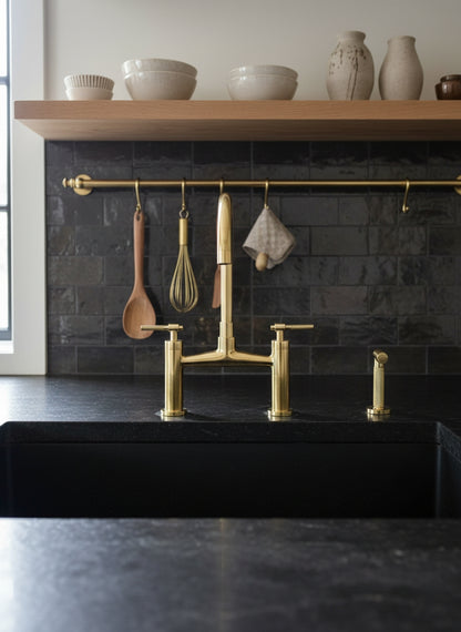 Unlacquered brass bridge kitchen faucet with matching side sprayer installed on a black sink with dark subway tile backsplash and brass hanging rail.