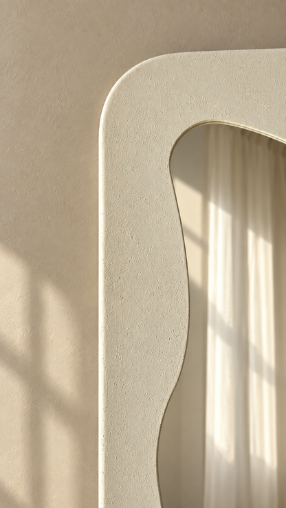 A close-up of a textured cream-colored wall mirror with a wavy, organic border, reflecting soft window light and curtains against a beige wall.