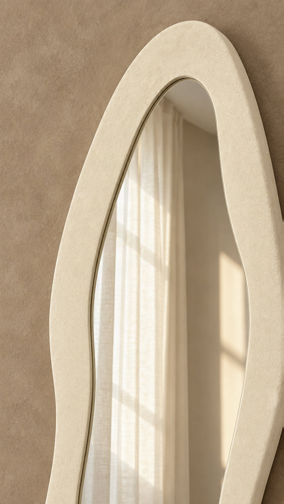 An organic-shaped cream-colored wall mirror with a thick textured frame, shown reflecting warm sunlight and curtains against a textured beige wall.