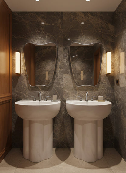 Organic brass wall mirrors above double pedestal sinks in marble bathroom.