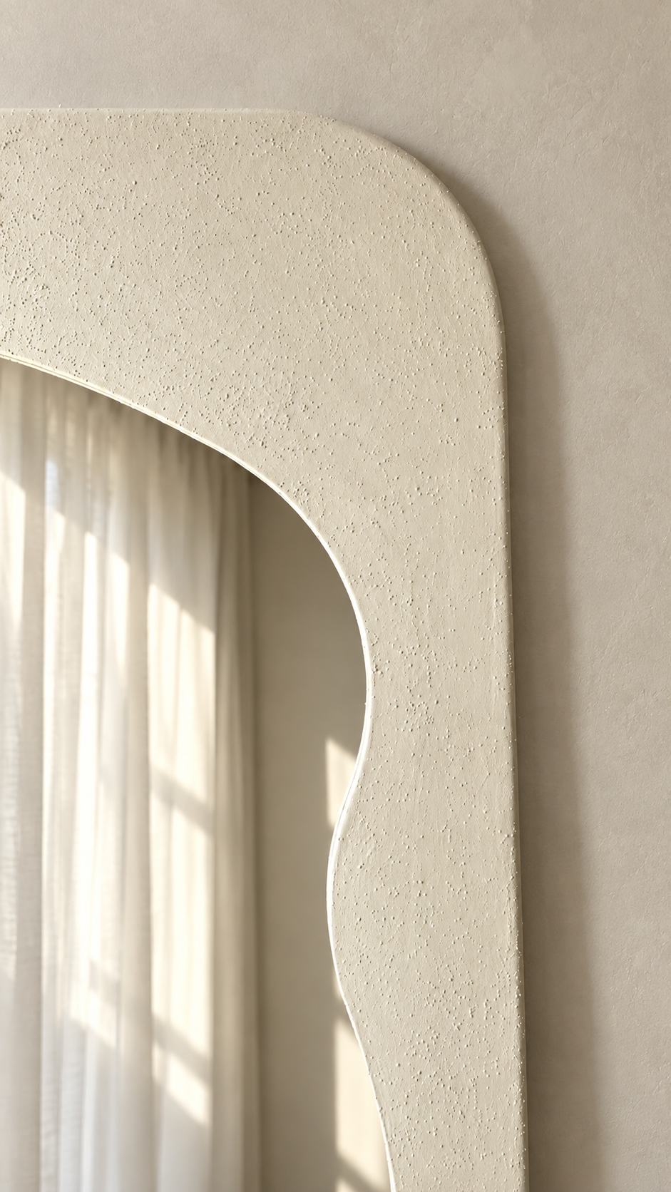 A close-up of a textured cream-colored wall mirror with a curved, scalloped border, reflecting soft sunlight and white curtains against a neutral beige wall.