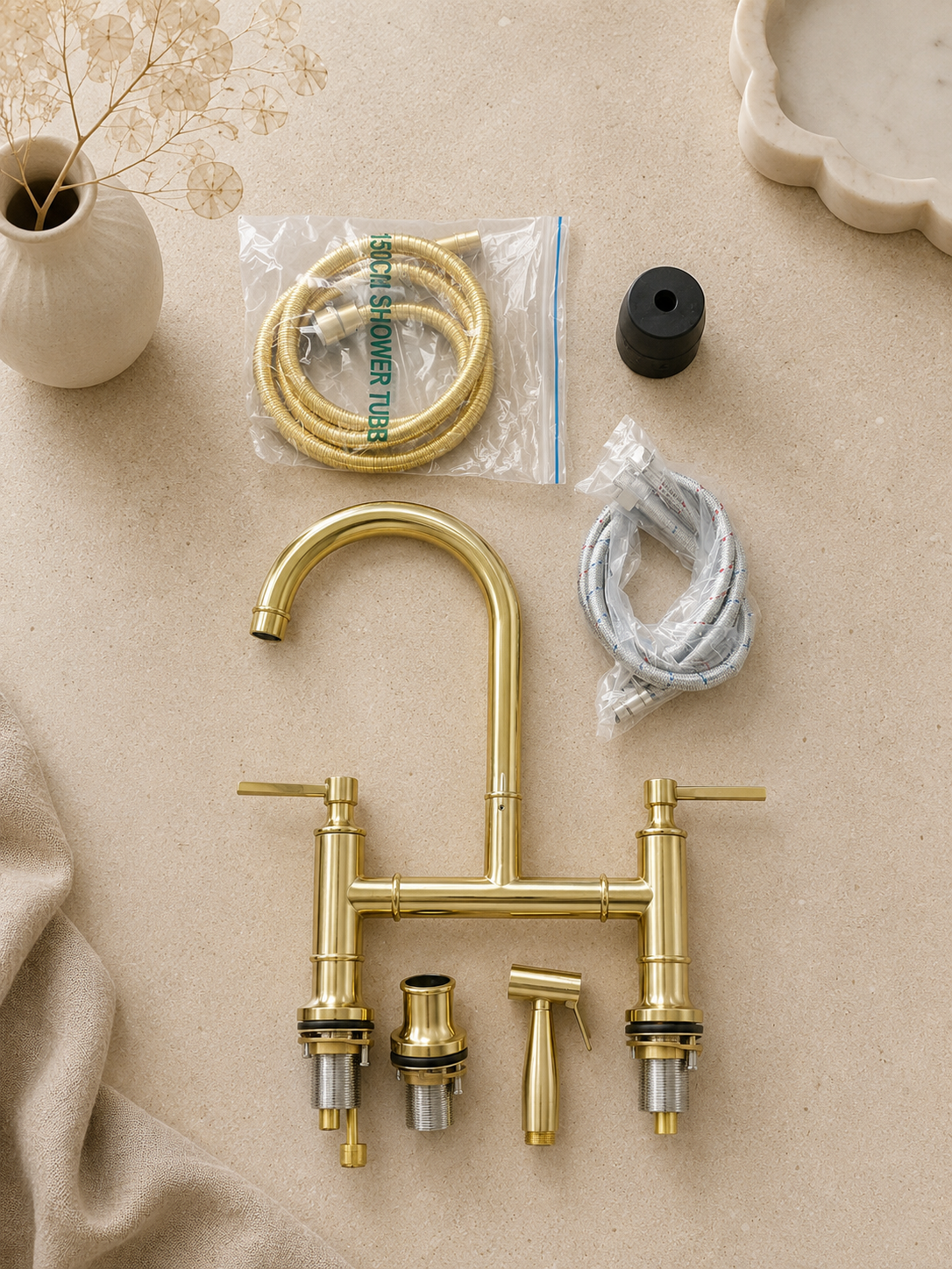 A brass double sink faucet set laid out on a beige surface, including the faucet body, two handles, a sprayer, a bag of hose, a black cap, and a wrapped flexible tube. Decorative props include a small vase with dried stems and a cream dish in the background.