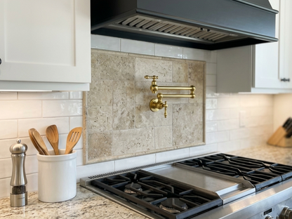 Unlacquered brass wall-mounted pot filler faucet installed above a professional gas range with a natural stone tile backsplash in a luxury kitchen.