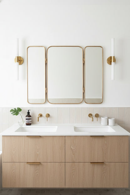 Three-panel brass mirror hung above a double sink vanity with white and light wood décor.