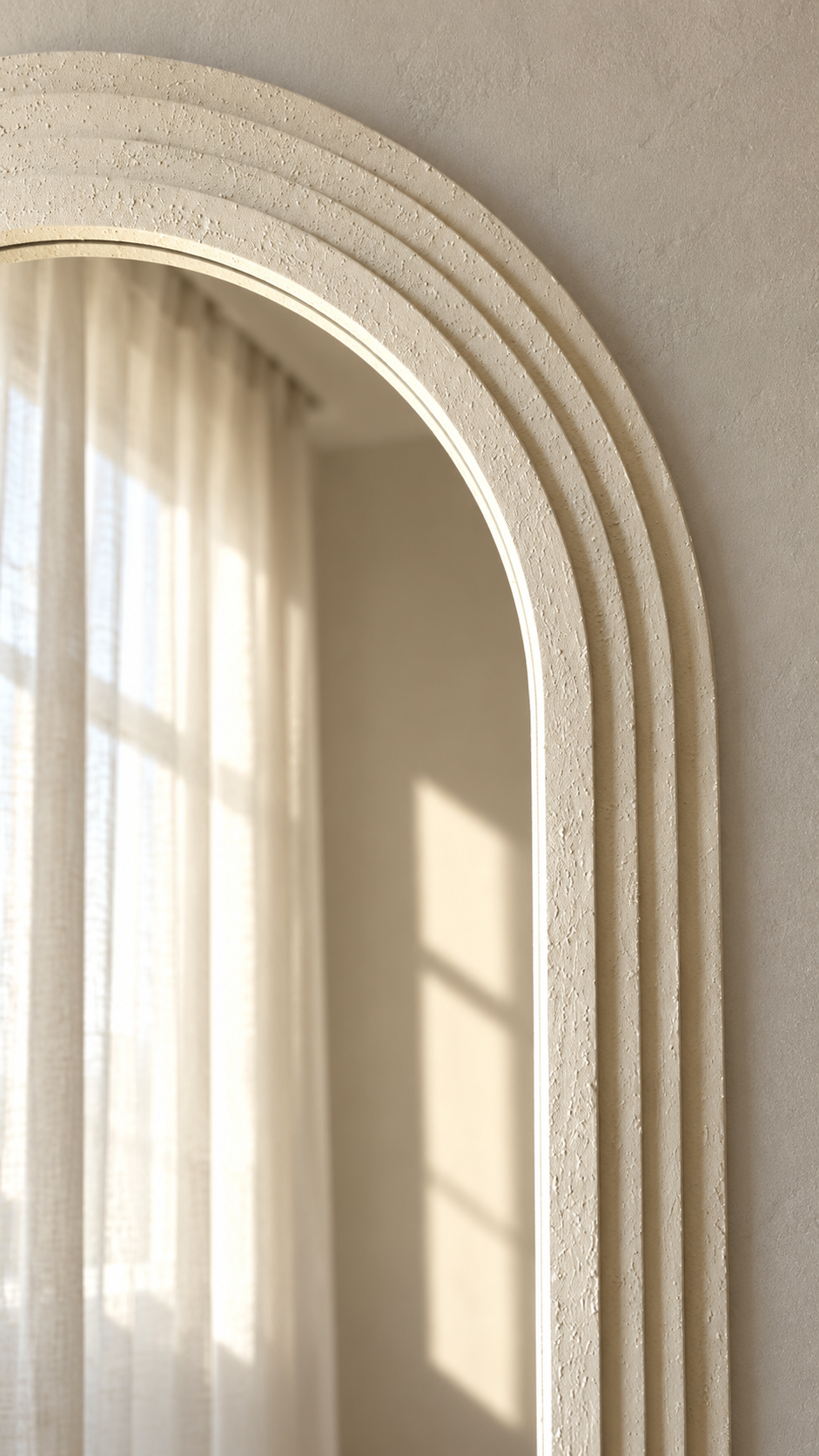 An arched decorative wall mirror with a textured cream-colored frame, mounted on a matching textured beige wall. The reflection shows a soft-focus interior with sheer curtains and warm sunlight.