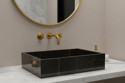 Black marble rectangular vessel sink with gold-tone wall faucet and round mirror.