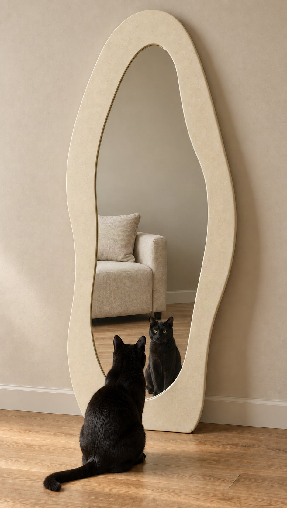 An organic-shaped full-length floor mirror with a cream-colored frame, leaning against a beige wall on a wooden floor. A black cat sits in front of the mirror, looking at its reflection.