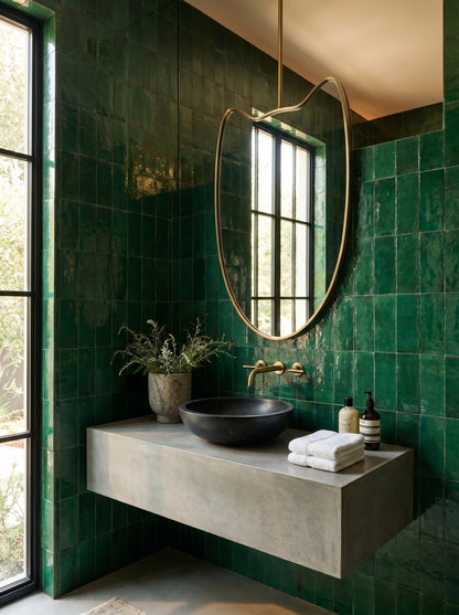 Organic brass hanging mirror above a floating concrete vanity with emerald green zellige tiles.