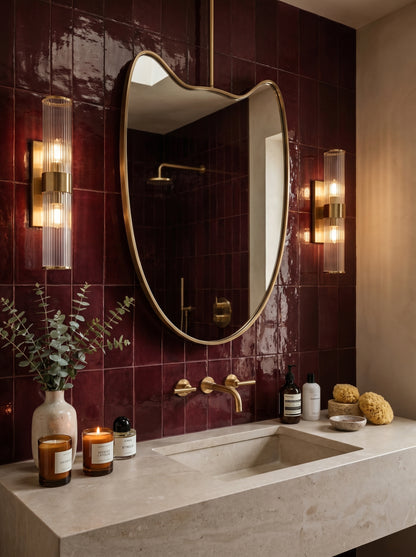 Wavy brass suspended mirror above a stone vanity with burgundy zellige tiles and brass sconces.