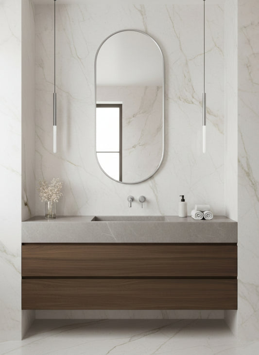 Silver capsule mirror above wood floating vanity in marble bathroom.