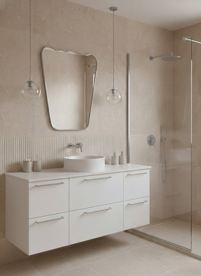 Silver asymmetrical brass mirror above white floating vanity in beige bathroom.