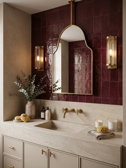 Ceiling-mounted brass arch mirror styled over a stone vanity with deep burgundy tile walls and brass sconces.