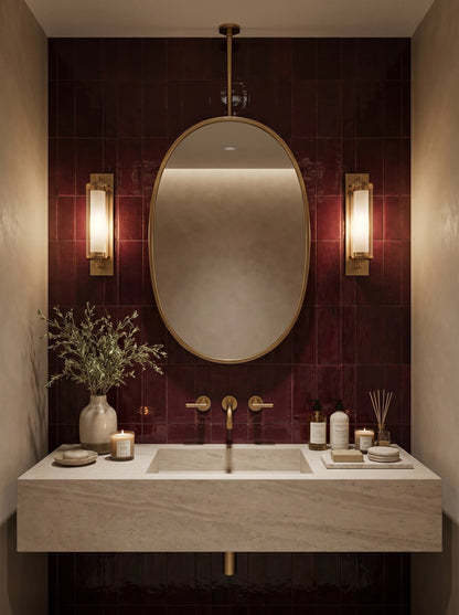 Suspended oval brass mirror above a stone vanity with burgundy zellige tiles and wall-mounted brass faucet.