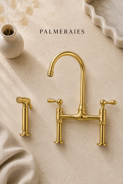 A polished brass bridge faucet set with a high-arc spout, two lever handles, and a matching side sprayer, arranged on a warm beige stone surface with decorative props. The brand name PALMERAIES is printed at the top.