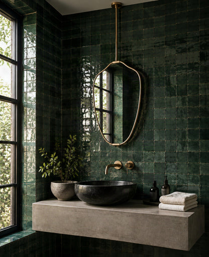 A modern bathroom vanity featuring dark green glossy tile walls, a floating concrete sink countertop, a black vessel sink, and an organic-shaped brass mirror.